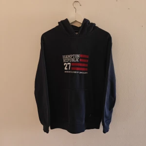 Hampton Republic hoodie - Mörkblå hoodie från Hampton Republic med tryck framtill i vitt och rött. Klassisk känguruficka och huva. Tillverkad i mjuk bomullsblandning (80% bomull, 20% polyester). Perfekt för chill dagar eller när du vill ha en enkel och snygg look.