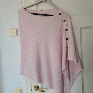 Ljusrosa stickad poncho Hampton Republic - Superfin ljusrosa stickad poncho från Hampton Republic med snygg struktur och dekorativa knappar längs ena axeln. Tröjan har en lös passform och är perfekt för lager-på-lager. Passar dig som gillar en chill och stilren look.