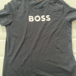 Svart BOSS t-shirt med tryck - Svart t-shirt från BOSS med klassisk rund hals och vit BOSS-logga framtill. T-shirten är kortärmad och har en enkel, stilren design som passar perfekt till jeans eller shorts. Tillverkad i mjuk bomull för en skön känsla hela dagen. Köpt på room1007