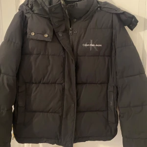 Svart pufferjacka Calvin Klein - Svart pufferjacka från Calvin Klein Jeans med broderad logga på bröstet. Jackan har hög krage, dragkedja och tryckknappar framtill samt en mysig fleecefodrad insida. Perfekt för kalla dagar och riktigt snygg. Väldigt varm. Passar både kille/tjej. Använd fåtal gånger. 700kr men pris kan diskuteras vid snabb affär