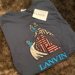 Lanvin X Fila T-shirt - Säljer denna mörkblå t-shirten från Lanvins collab med fila för riktigt bra pris. T-shirten har ett unikt broderat mönster på framsidan. T-shirten har en rund halsringning och är kortärmad. T-shirten är i storlek S och har passformen slim. Denna T-shirten är perfekt för dig som vill ha något stilrent och unikt till sommarn. 