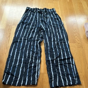 Randiga palazzo byxor från H&M 38 - Snygga randiga ankellånga byxor från H&M i svart och blått med vita ränder. Byxorna har en bekväm loose fit och är försedda med snörning i midjan för en justerbar passform. Perfekta för en avslappnad stil.