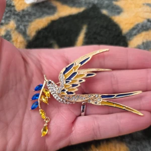 Färgglad fågelbrosch med strass - Snygg brosch i form av en fågel med glittrande strassdetaljer. Fjädrarna är i blått och gult, och fågeln håller en kvist med färgglada stenar. Perfekt för att liva upp vilken outfit som helst!