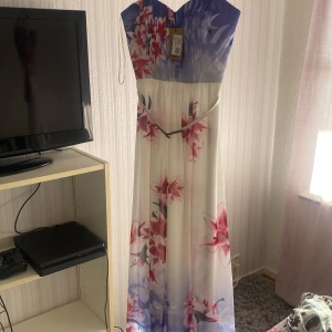 Blommig långklänning från Jane Norman - Säljer en vacker långklänning från Jane Norman med blommigt mönster i rosa och lila nyanser. Klänningen har en v-ringad överdel och ett elegant bälte i midjan. Perfekt för sommarens festligheter!