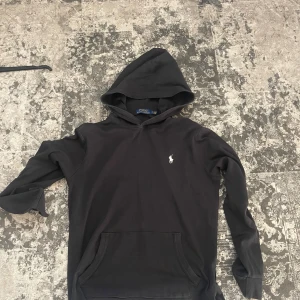 Svart hoodie från Polo Ralph Lauren - Säljer en stilren svart hoodie från Polo Ralph Lauren med det klassiska logotypbroderiet på bröstet. Tröjan har en praktisk känguruficka och en bekväm huva. Perfekt för en avslappnad look.