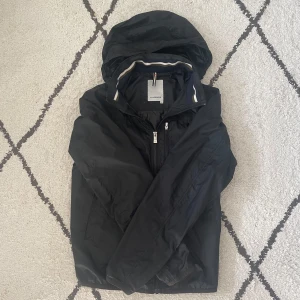 Svart vindjacka från J.Lindeberg - Säljer en stilren svart vindjacka från J.Lindeberg. Jackan har en praktisk dragkedja framtill och en justerbar huva. Perfekt för blåsiga dagar. Passar bra till både vardag och träning.