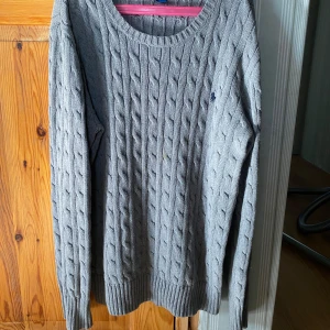 Grå stickad tröja från Ralph Lauren - Säljer en stilren grå stickad tröja från Ralph Lauren med kabelstickat mönster. Tröjan har långa ärmar och en rund halsringning. Perfekt för kyligare dagar.