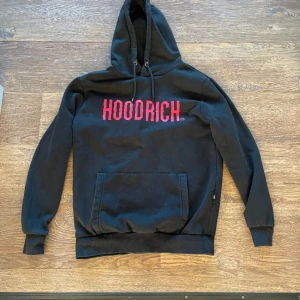 Svart hoodie från Hoodrich - Säljer en svart hoodie från Hoodrich med röd text på bröstet. Den har en klassisk känguruficka och justerbar huva med snören. Perfekt för en avslappnad.