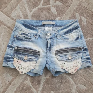 Blå jeansshorts från Nitty Lux - Snygga blå jeansshorts från Nitty Lux med coola detaljer som nitar och dragkedjor på fickorna. Perfekta för en trendig look i sommar. Shortsen har en klassisk femficksdesign och en ljus tvätt för en avslappnad stil.
