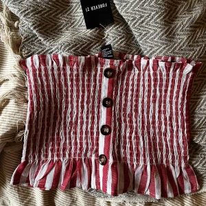 Randig tubtopp från Forever 21 - Säljer en randig tubtopp från Forever 21 i rött och vitt. Toppen har knappar framtill och en volangdetalj vid över- och nederkant. Aldrig använd. 