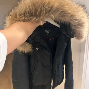 Svart dunjacka med päls från Hollies - Snygg svart dunjacka från Hollies med en stor, fluffig päls på huvan. Jackan har dragkedja och knappar framtill samt praktiska fickor.