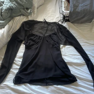 Svart långärmad topp med öppen rygg - Snygg svart långärmad topp med transparenta ärmar och en elegant öppen rygg. Toppen har en unik design med knytning i ryggen och en smickrande passform. Perfekt för en kväll ute eller en speciell tillställning. Säljer den för den sitter lite tajt på mig