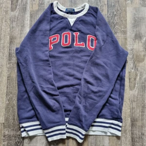 Blå tröja från Ralph Lauren - Säljer en blå tröja från Ralph Lauren med texten 'POLO' i rött och vitt på framsidan. Tröjan har långa ärmar och ribbade muddar i grått och vitt vid ärmslut och nederkant. Perfekt för en avslappnad stil.