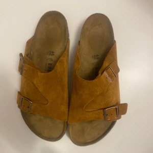 Bruna sandaler från Birkenstock - Säljer ett par bruna sandaler från Birkenstock i mocka med justerbara spännen. Perfekta för sommarens varma dagar. Sandalerna har en bekväm fotbädd och en slitstark sula.