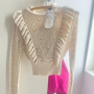 Beige stickad tröja med volang och pärlor - Säljer en beige stickad tröja med vackra volangdetaljer och pärlor framtill. Tröjan har ett fint mönster och långa ärmar.💕 Passar utmärkt till både vardag och festliga tillfällen.💕💕 Tröjan är oanvänd och i nyskick🎂