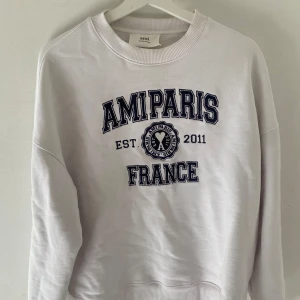 Vit sweatshirt från Ami Paris - Säljer en stilren vit sweatshirt från Ami Paris med tryck framtill och en liten logga bak i nacken 