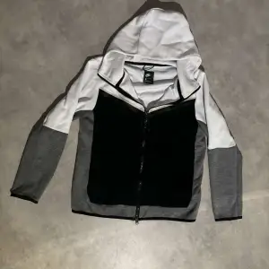 Säljer en stilren huvtröja från Nike i svart, vit och grå. Tröjan har en dragkedja framtill och en praktisk huva. Perfekt för en sportig look eller avslappnade dagar. Passar även S.