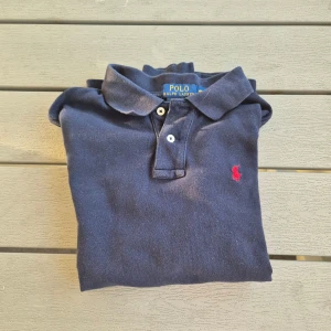 Blå pikétröja från Ralph Lauren - Säljer en klassisk blå pikétröja från Ralph Lauren med lång ärm och röd broderad logga på bröstet. Tröjan har en krage med knappar och är perfekt för en stilren look. Passar både till vardags och mer uppklädda tillfällen.