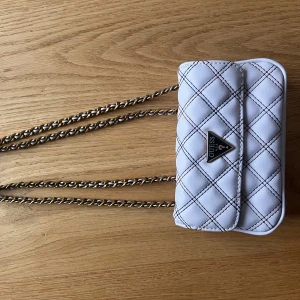 Vit quiltad axelväska från Guess - Snygg vit quiltad axelväska från Guess med gysterbar kedjeaxelrem så att man kan ha den crossbody. Med ett elegant rutmönster. Väskan har en praktisk storlek och stängs med en tryckknapp. Den är använd några gånger men är i väldigt gott skick utan tecken på användning.