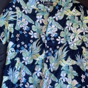 Blommig kortärmad skjorta från Raintree Republic - Säljer en färgglad kortärmad skjorta från Raintree Republic med tropiskt blommönster i blått, grönt och vitt. Perfekt för sommarens äventyr! 🌺