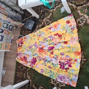 Gul blommig klänning - Säljer en färgglad gul klänning med blommigt mönster i lila, rosa och grönt. Klänningen har en axelbandslös design och är perfekt för sommarens festligheter. Den har en luftig passform och är gjord i ett lätt material.