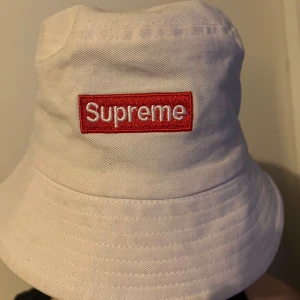 Vit bucket hat från Supreme - Snygg vit bucket hat från Supreme med det ikoniska röda och vita logotypmärket framtill. 