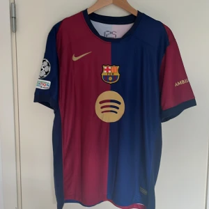 FC Barcelona fotbollströja 24/25 Lamine Yamal - Säljer en officiell FC Barcelona fotbollströja från Nike. Tröjan är i de klassiska färgerna röd och blå med klubbens emblem på bröstet. Den har korta ärmar och trycket 'Lamine Yamal 19' på ryggen. UEFA Champions League-märke på ärmen.