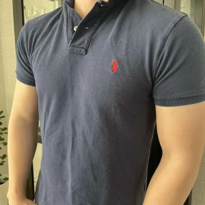 Blå pikétröja från Ralph Lauren - Säljer en stilren blå pikétröja från Ralph Lauren med röd broderad logga på bröstet. Tröjan har en klassisk krage med knappar. Perfekt för en avslappnad men ändå snygg look, säljes då den är för liten för mig. Väldigt bra skick på piken.