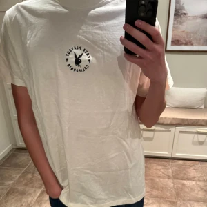 Vit t-shirt med tryck från Playboy - Säljer en vit t-shirt från Playboy med ett litet tryck på framsidan och ett större färgglatt tryck på baksidan. T-shirten har en normal passform och korta ärmar. Perfekt för en avslappnad stil!