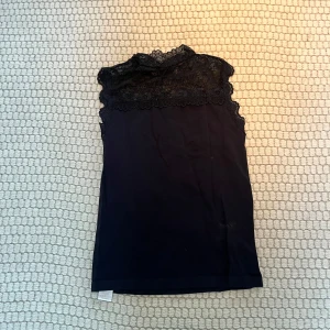 Svart ärmlös topp med spetsdetaljer - Snygg svart ärmlös topp med vacker spetsdetalj vid halsen. Perfekt för en elegant look. Toppen har en tight passform och är tillverkad i ett mjukt material. S/M