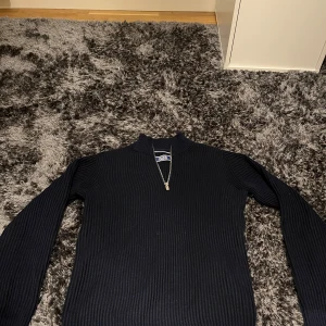 Svart ribbad tröja från Jack & Jones - Säljer en stilren svart ribbad tröja från Jack & Jones. Tröjan har en dragkedja vid halsen och långa ärmar, perfekt för kyligare dagar. Passar både till vardags och mer uppklädda tillfällen. Hör av dig vid intresse!