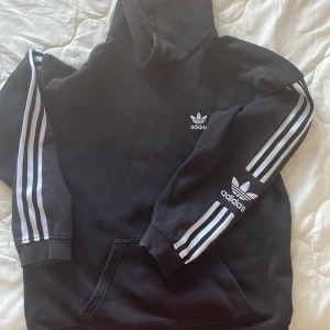 Svart hoodie från Adidas - Säljer en svart hoodie från Adidas med de klassiska tre ränderna i vitt längs ärmarna. Den har en känguruficka och Adidas-loggan på bröstet och ärmen. Perfekt för en sportig look eller avslappnade dagar.