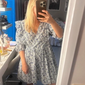 Blommig klänning med volangdetaljer - Söt blommig klänning i blått och vitt med volangdetaljer på axlarna. Klänningen har en omlott likande och trekvartsärmar, perfekt för vår och sommar. Använd fåtal gånger från hm, väldigt stor i storleken 