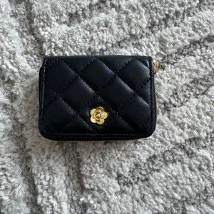 Svart quiltad plånbok med blomdetalj - Snygg svart quiltad plånbok med en elegant guldfärgad blomdetalj på framsidan. Plånboken har en praktisk dragkedja i guld och flera fack för kort och kontanter. Perfekt för att hålla ordning på dina tillhörigheter med stil.