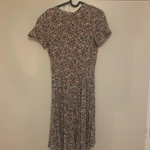Blommig klänning med korta ärmar - Säljer en söt blommig klänning med korta ärmar och dragkedja i ryggen. Klänningen har ett färgglatt mönster i rosa, beige och svart. Perfekt för vår och sommar! Hella känning är 52 cm lång så den räcker ner till knät.