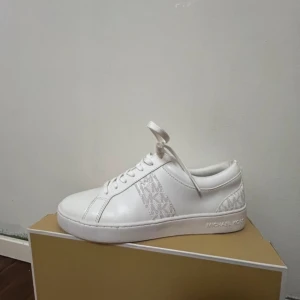 Vita sneakers från Michael Kors - Snygga vita sneakers från Michael Kors med diskret logotypmönster på sidorna. Skorna har en klassisk design med snörning och rund tå, perfekt för en stilren look. Tillverkade i syntetmaterial för en modern känsla.