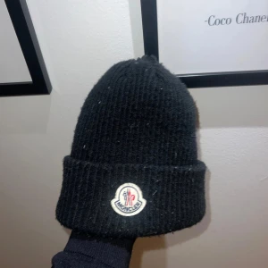Svart ribbad mössa från Moncler - Snygg svart ribbad mössa från Moncler med deras ikoniska logga framtill. Perfekt för att hålla värmen under kyliga dagar. Enkel och stilren design som passar till det mesta.