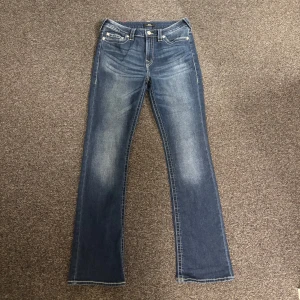 Blå jeans från True Religion - Snygga blå jeans från True Religion med klassisk femficksdesign och broderade bakfickor. De har en bootcut-stil och är perfekta för en avslappnad look. Märkeslappen på baksidan ger en extra touch av stil.
