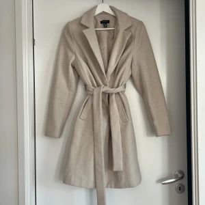 Beige kappa från New Look - Säljer en stilren beige kappa från New Look. Den har ett bälte i midjan och långa ärmar, perfekt för höst och vår. Använd en gång. 