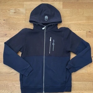 Svart huvtröja från Sail Racing - Snygg svart huvtröja från Sail Racing med dragkedja och en praktisk bröstficka. Tröjan har en broderad logga på bröstet och huvan, samt ribbade muddar vid ärmslut och nederkant. Perfekt för kyliga dagar!