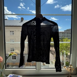 Svart meshtröja med hög krage - Säljer en svart långärmad meshtröja med hög krage. Tröjan är genomskinlig och har ett subtilt mönster i tyget som ger en cool look. Perfekt att styla lager på lager eller som statement-piece.