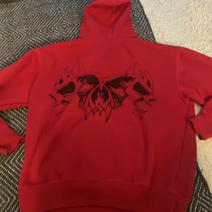 Röd hoodie med döskalletryck från D-Way - Säljer en röd hoodie från D-Way med ett stort svart tryck av två döskallar och en fjäril på ryggen. Tröjan har huva och ribbade muddar. Perfekt för dig som gillar unika och lite edgy plagg!