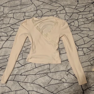 Beige omlottopp med spets från Dazy - Säljer en beige omlottopp från Dazy med långa ärmar och fina spetsdetaljer längs den v-ringade omlottringningen. Toppen har en figurnära passform och är tillverkad i ett mjukt, stretchigt material.