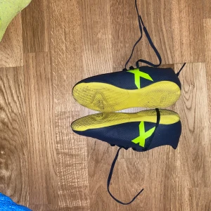Adidas fotbollsskor i blått och gult - Säljer ett par blå fotbollsskor från Adidas med gula detaljer och snörning. Skorna har platt sula och är perfekta för inomhusspel. Adidas-loggan syns tydligt på sidan och sulan är gul.perfekta för inomhusspel eller grusspel perfekta för din son eller dotter som vill prova på fotboll eller behöver skor till gympan på skolan