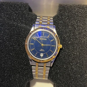 SILVAS herrklocka med blå urtavla - Stilren herrklocka från SILVAS med blå urtavla och datumvisning. Klockan är i silverfärgad metall med guldfärgade detaljer. Klassisk design som passar till många tillfällen.