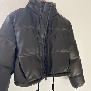 Svart/brun pufferjacka i skinnimitation -  svart/brun pufferjacka i skinnimitation med hög krage och dragkedja framtill. Jackan är croppad och har justerbar dragsko i nederkant. Perfekt för dig som vill ha en trendig och varm jacka med cool attityd. Aldrig använd storlek s 