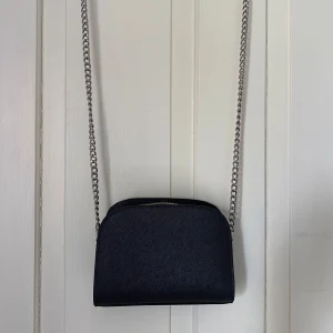Svart axelväska med kedjerem från H&M - Säljer en stilren svart axelväska från H&M med silverfärgad kedjerem och dragkedja upptill. Väskan har en enkel, rundad form och är perfekt för att bära det viktigaste. Insidan är ljus färgad. > Man kan ha med det mesta som en telefon, nycklar, smink ( mensskydd ) osv!