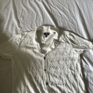 Vit stickad pike från H&M - Vit pike från H&M med stickat diamantmönster och svarta knappar. Tröjan har korta ärmar och en klassisk krage, perfekt för en clean och stilren look. Perfekt på varma sommardagar eftersom den är så luftig och skön, riktigt snygg också men för liten för mig