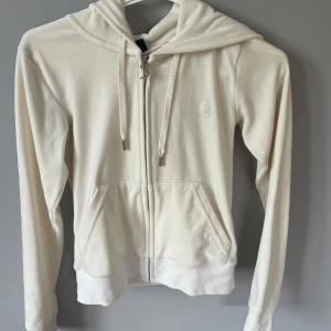 Vit hoodie med dragkedja från Juicy Couture - Säljer en klassisk krämvit hoodie från Juicy Couture i mjukt material. Jackan har dragkedja framtill, huva med snören och fickor på magen. Liten broderad logga på bröstet. Perfekt för en avslappnad och trendig look.