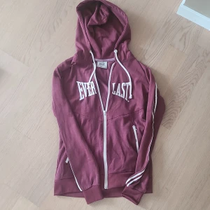 Vinröd hoodie från Everlast - Säljer en vinröd hoodie från Everlast med vit dragkedja och vita detaljer. Tröjan har huva, långa ärmar med ränder på sidorna och fickor framtill. Perfekt för en sportig och avslappnad stil. Inga tecken på slitage, kom privat för fler bilder eller mått!